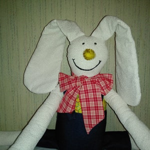 May include: A handmade white bunny doll with long floppy ears, a yellow nose, and a stitched smile. It wears a red and white plaid bow tie and a blue and yellow outfit. A charming, whimsical stuffed animal.