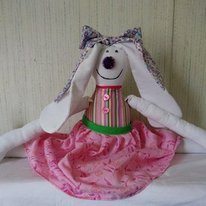 May include: A white fabric bunny doll with long ears, wearing a pink dress with floral print and a green sash. The doll has a purple glitter nose and pink buttons on its striped bodice.