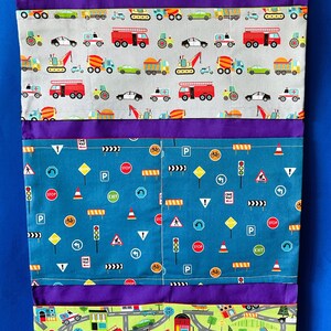 May include: A purple fabric storage organizer with three pockets. The top pocket has a gray fabric with a repeating pattern of various vehicles. The middle pocket has a blue fabric with a repeating pattern of traffic signs and symbols. The bottom pocket has a green fabric with a repeating pattern of a city street scene with vehicles and buildings.