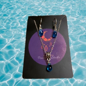 Könnte beinhalten: Ein silbernes Halsketten- und Ohrring-Set mit blauen, tropfenförmigen Glasperlen. Die Halskette hat einen silbernen Anhänger mit einem komplizierten Design. Das Set wird auf einem violetten Hintergrund mit dem Text "Pixie Studio" präsentiert.