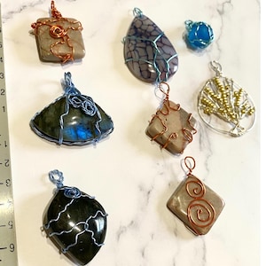 May include: Assortment of wire-wrapped pendants featuring various stones and designs. The pendants include a square stone with copper wire, a teardrop-shaped stone with blue wire, a fan-shaped stone with blue wire, a square stone with copper wire, a tree-shaped design with gold wire, and a round stone with blue wire.