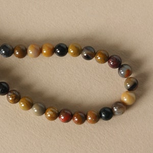 May include: A necklace of polished gemstone beads in various earth tones. The beads are round and have a glossy finish, with colours ranging from black and brown to tan, red, and grey. The beads are strung together, forming a circular shape.