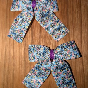 May include: Two blue and pink hair bows with a purple centre. The bows have a pattern of small blue, pink, and yellow owls.