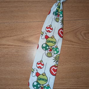 Puede incluir: Una corbata blanca con un patrón repetido de adornos navideños y el Grinch. Los adornos son rojos, verdes y blancos, con la cara del Grinch en algunos. La corbata tiene una banda elástica negra.