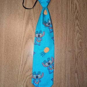 May include: A turquoise tie featuring a repeating pattern of a cartoon character wearing sunglasses and a lei, along with pineapples. The tie has a black elastic band and is made of fabric.