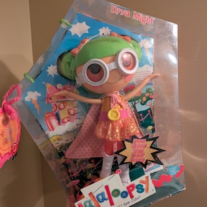 May include: A Lalaloopsy Dyna Might doll in original packaging. The doll has green hair, white goggles, a yellow star-patterned dress with a pink belt and button, a pink cape, and white boots. The box features the text Dyna Might, Lalaloopsy, and Pow Bang Zas Paff Pang.