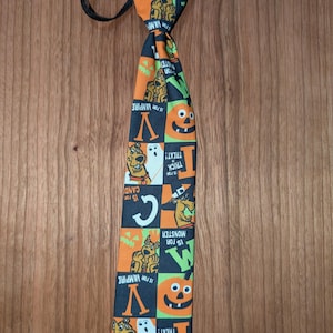 Może przedstawiać: Krawat o tematyce Halloween z czarnym tłem i pomarańczowymi i zielonymi kwadratami. Krawat przedstawia postacie z kreskówek, dynie, duchy oraz frazy "V is for Vampire", "Trick or Treat!" i "C is for Candy".