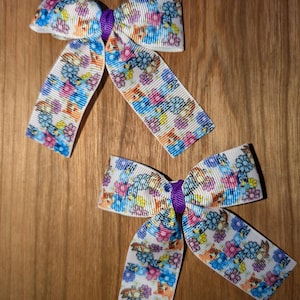 May include: Two white hair bows with a purple centre. The bows are made of ribbon with a colourful print of cartoon animals and flowers.