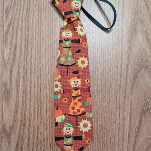 May include: A novelty tie featuring a repeating pattern of scarecrows, sunflowers, and pumpkins on a rust-colored background. The tie has a black elastic band for easy wear. The scarecrows are dressed in colorful outfits.