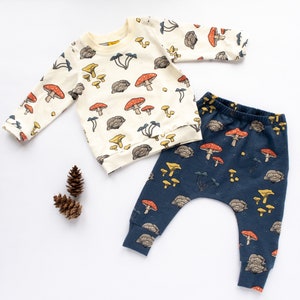 May include: A cream-colored long-sleeved sweatshirt and navy blue pants set for babies, adorned with a whimsical mushroom pattern in red, yellow, and brown. The set is made of soft fabric and is perfect for a baby.