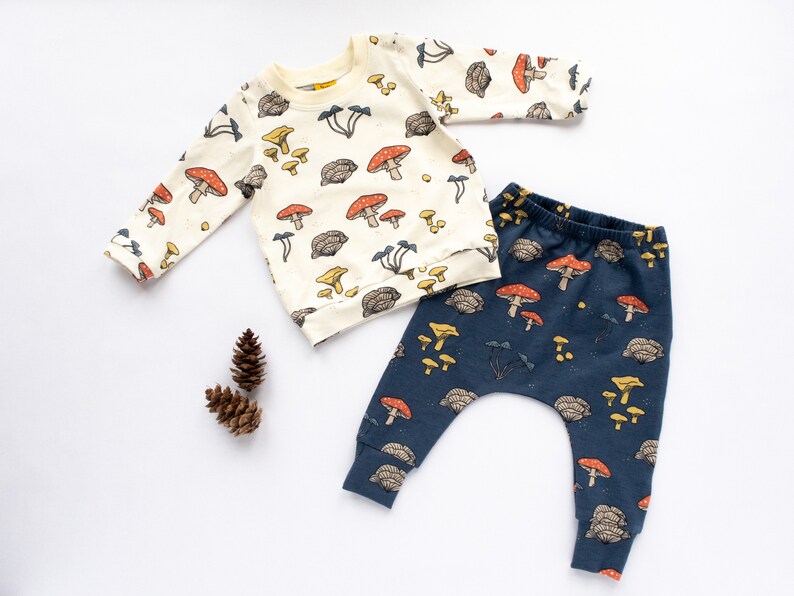 May include: A cream-colored long-sleeved sweatshirt and navy blue pants set for babies, adorned with a whimsical mushroom pattern in red, yellow, and brown. The set is made of soft fabric and is perfect for a baby.