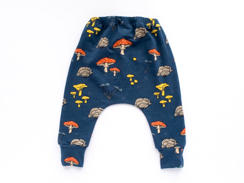 May include: Dark blue baby pants with a whimsical mushroom pattern. The design features various mushroom illustrations in shades of red, yellow, and beige. The pants have a comfortable, elastic waistband and cuffs.