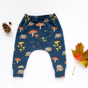 May include: Dark blue baby pants with a whimsical mushroom pattern in orange, yellow, and beige. The pants have a comfortable, relaxed fit and are made of soft fabric. A pinecone and leaf are next to the pants.