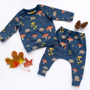 May include: A navy blue baby outfit set featuring a long-sleeved sweatshirt and pants. The set is patterned with red, yellow, and brown mushrooms, along with other forest elements. The outfit is made of soft fabric.