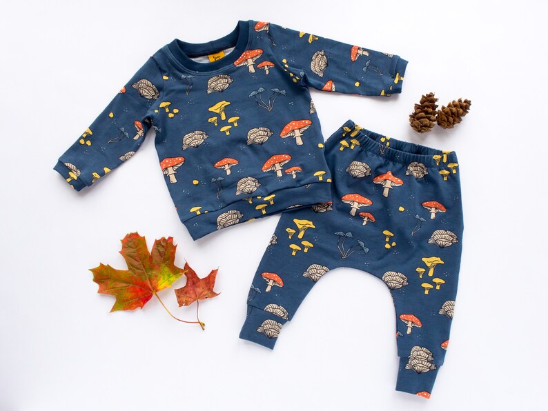 May include: A navy blue baby outfit set featuring a long-sleeved sweatshirt and pants. The set is patterned with red, yellow, and brown mushrooms, along with other forest elements. The outfit is made of soft fabric.