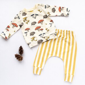 May include: A baby outfit set with a long-sleeved white shirt with a mushroom print and yellow and white striped pants.
