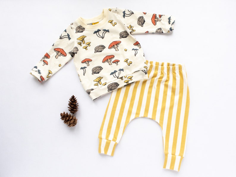 May include: A baby outfit set with a long-sleeved white shirt with a mushroom print and yellow and white striped pants.