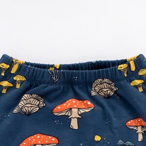 May include: Dark blue elastic waistband pants with a repeating pattern of orange and white mushrooms, yellow mushrooms, and small white dots. The waistband is lined with yellow fabric.