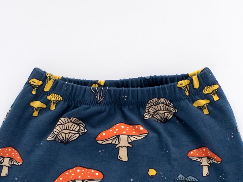 May include: Dark blue elastic waistband pants with a repeating pattern of orange and white mushrooms, yellow mushrooms, and small white dots. The waistband is lined with yellow fabric.