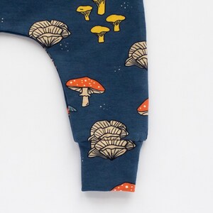 May include: Close-up of a navy blue baby pant leg with a repeating mushroom pattern. The mushrooms are in shades of orange, yellow, and beige. The cuff is a solid navy blue.