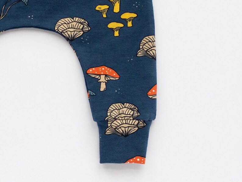 May include: Close-up of a navy blue baby pant leg with a repeating mushroom pattern. The mushrooms are in shades of orange, yellow, and beige. The cuff is a solid navy blue.