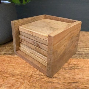 May include: A set of six wooden coasters in a wooden holder. The coasters are stacked and the holder is made of light brown wood.