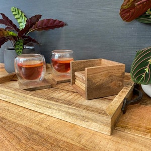 May include: A wooden tray with two glasses of tea and a stack of wooden coasters. The tray has a natural wood finish and a metal handle. The glasses are clear and filled with a reddish-brown liquid. The coasters are square and have a natural wood finish.