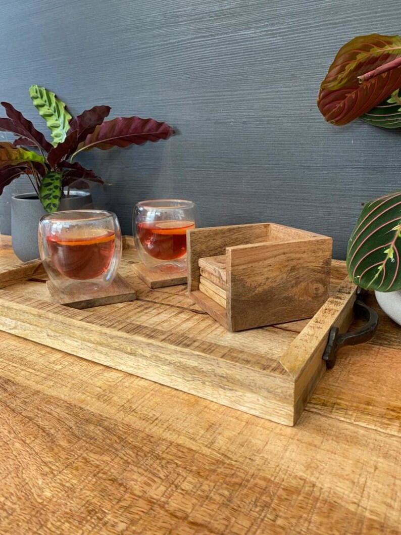 May include: A wooden tray with two glasses of tea and a stack of wooden coasters. The tray has a natural wood finish and a metal handle. The glasses are clear and filled with a reddish-brown liquid. The coasters are square and have a natural wood finish.