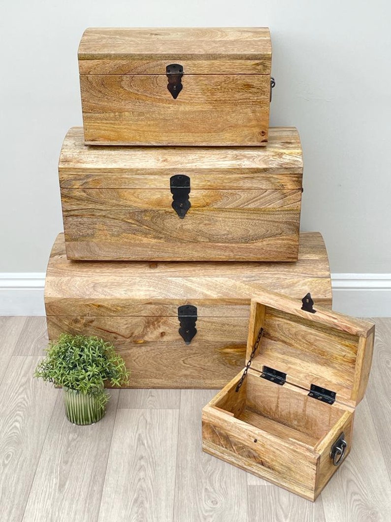 May include: Set of three wooden storage chests with arched lids and black metal hardware. The chests are various sizes, with the smallest one open, revealing the interior. The wood has a natural grain and warm tones.
