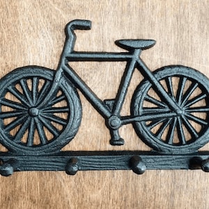 May include: Black cast iron bicycle-shaped wall hook with three hooks. The bicycle design features detailed wheels, frame, and handlebars. The hook is mounted on a wooden surface, suitable for hanging keys, coats, or other items.