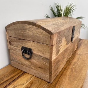 May include: A wooden chest with a curved top and metal hardware. The chest is made of light brown wood with visible grain and features a dark metal clasp and handle. The chest is sitting on a wooden surface.