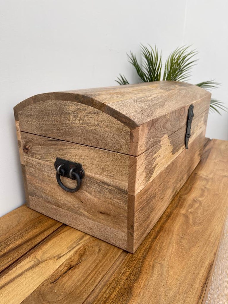 May include: A wooden chest with a curved top and metal hardware. The chest is made of light brown wood with visible grain and features a dark metal clasp and handle. The chest is sitting on a wooden surface.