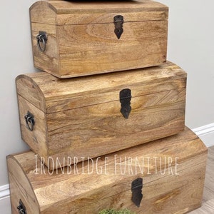 May include: Four wooden storage chests stacked, each with a curved top and metal hardware. The chests are various sizes, with the largest at the bottom. The wood has a warm, natural finish. The text "IRONBRIDGE FURNITURE" is visible.