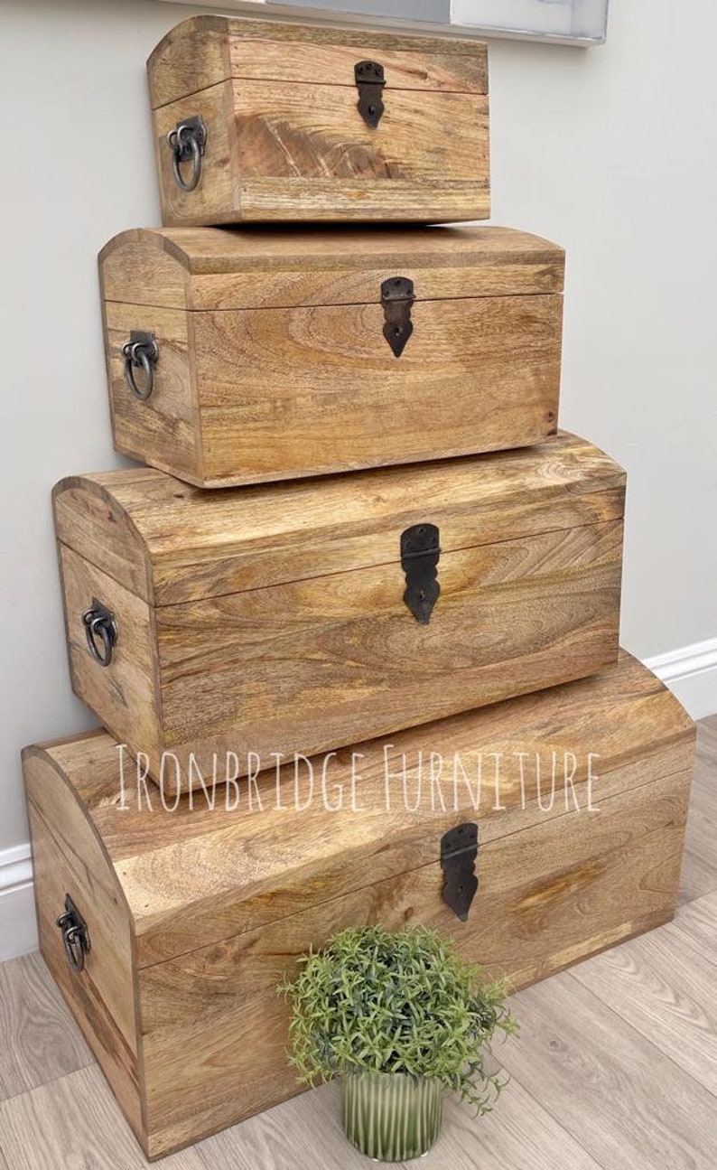 May include: Four wooden storage chests stacked, each with a curved top and metal hardware. The chests are various sizes, with the largest at the bottom. The wood has a warm, natural finish. The text "IRONBRIDGE FURNITURE" is visible.