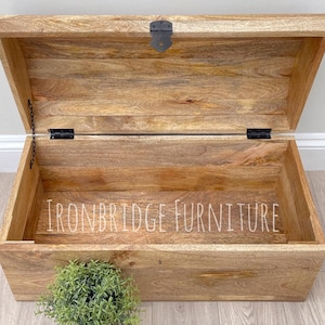 May include: Open wooden storage chest with a hinged lid and metal hardware. The chest is made of light brown wood with visible grain and the text "IRONBRIDGE FURNITURE" is visible inside the chest.