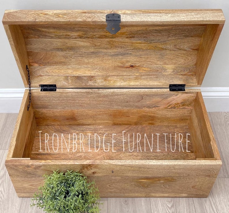 May include: Open wooden storage chest with a hinged lid and metal hardware. The chest is made of light brown wood with visible grain and the text "IRONBRIDGE FURNITURE" is visible inside the chest.