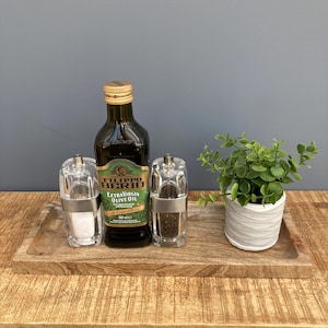 May include: A wooden tray with a bottle of Filippo Berio Extra Virgin Olive Oil and two glass salt and pepper shakers. The tray is on a wooden surface and there is a small potted plant in the background.