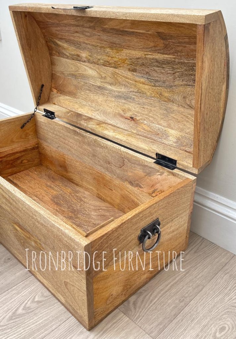 May include: Wooden storage chest with a curved lid, open to reveal the interior. The chest is made of light-colored wood with visible grain and features a metal handle and hinges. The text "IRONBRIDGE FURNITURE" is visible on the front.