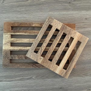 May include: Two wooden trivets with a natural finish. One is rectangular with horizontal slots, and the other is square with vertical slots. The trivets are designed to protect surfaces from heat.