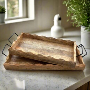 May include: Two rectangular wooden serving trays with wavy edges and black metal handles. The trays are made of light brown wood and are stacked on top of each other. The trays are on a white countertop.