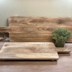 May include: Two wooden chopping boards, one larger than the other, with a natural wood finish. The larger board has a handle and the smaller board has a smaller handle. Both boards are sitting on a wooden surface.