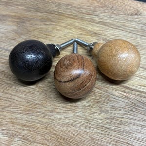May include: Three wooden cabinet knobs with metal screws. One knob is black, one is light brown, and the center knob is a medium brown with wood grain. The knobs are round and sit on a wooden surface.