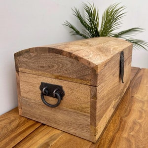 May include: A wooden chest with a curved top and metal hardware. The chest is made of light-colored wood with visible grain and features a metal handle and clasp. The chest is sitting on a wooden surface.