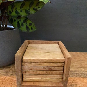 May include: A set of six wooden coasters in a wooden holder. The coasters are stacked on top of each other and are made of a light brown wood.