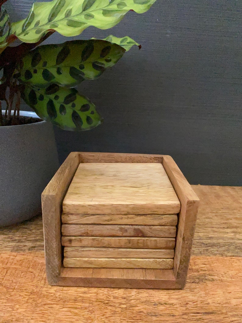 May include: A set of six wooden coasters in a wooden holder. The coasters are stacked on top of each other and are made of a light brown wood.