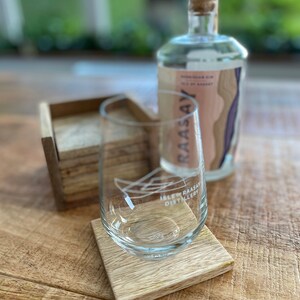 May include: A clear glass with the text "Isle of Raasay Distillery" on it, sitting on a wooden coaster. A clear glass bottle with a cork top and the text "Raasay" on it is in the background.