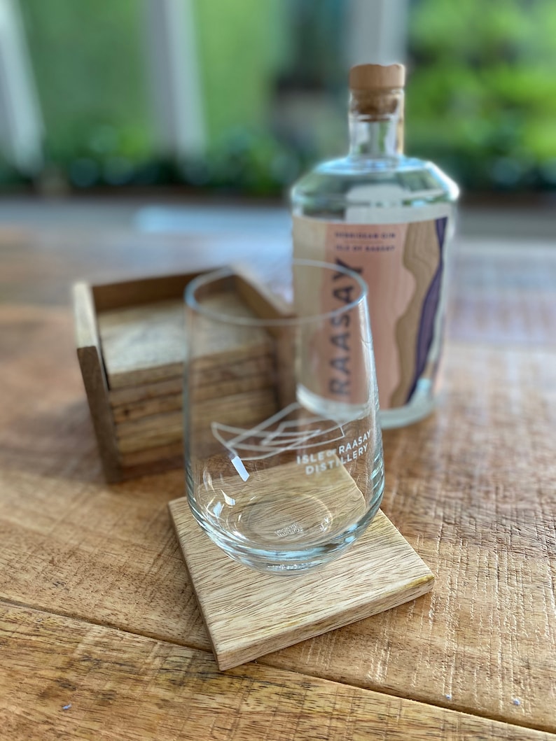 May include: A clear glass with the text "Isle of Raasay Distillery" on it, sitting on a wooden coaster. A clear glass bottle with a cork top and the text "Raasay" on it is in the background.