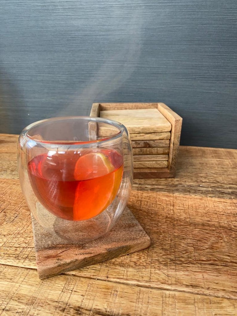 May include: A glass of red tea with a lemon slice in it, sitting on a wooden coaster. The tea is steaming and there is a stack of wooden coasters in the background.