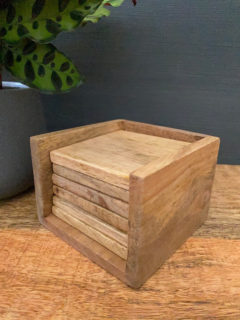 May include: A set of six wooden coasters in a wooden holder. The coasters are stacked and the holder is made of light brown wood.