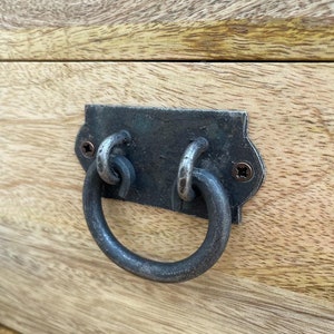 May include: Close-up of a wooden surface with a metal drawer pull. The pull is a dark, aged metal with a ring handle. The wood has a light, natural finish with visible grain.